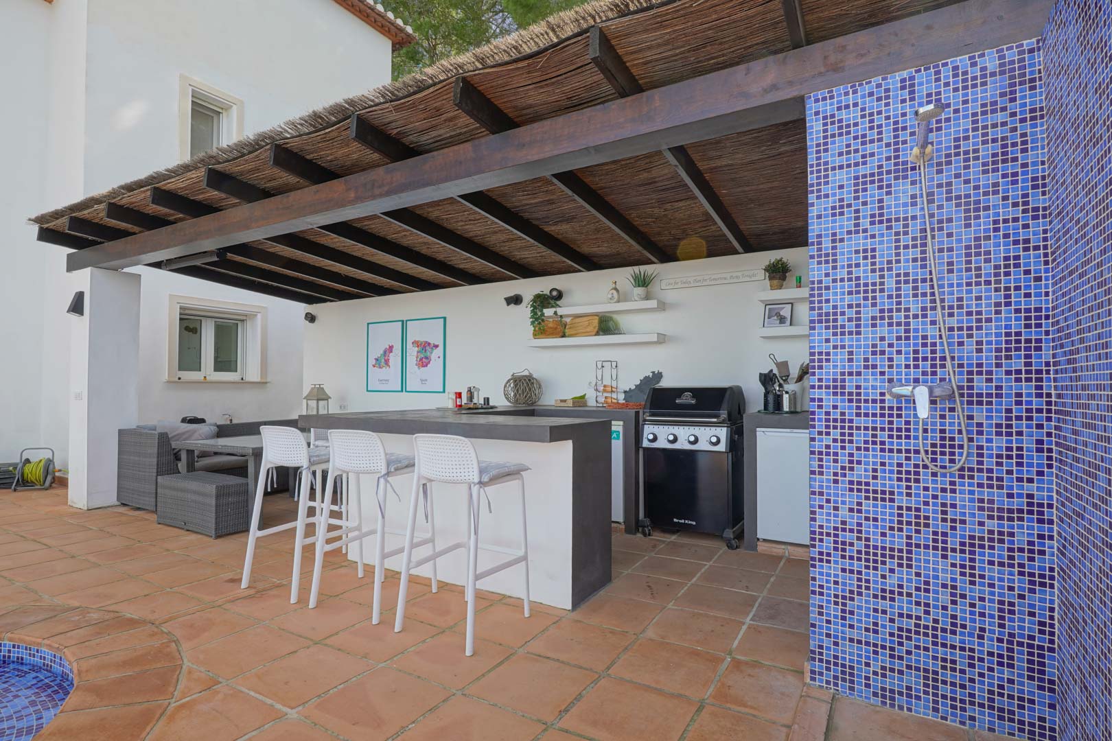 Außenküche unter einer Holzpergola mit Reetdach, ausgestattet mit Gasgrill, Bar mit Hockern und einer Außendusche mit blauen Mosaikfliesen.