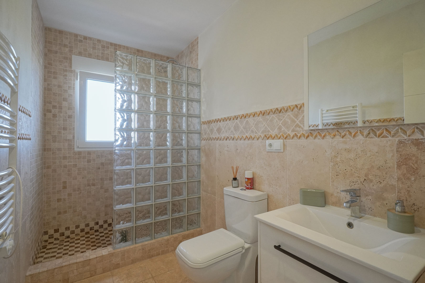 Badezimmer mit begehbarer Dusche, Glasbaustein-Trennwand, erdfarbenen Mosaikfliesen, weißem Waschtisch und Handtuchheizkörper.