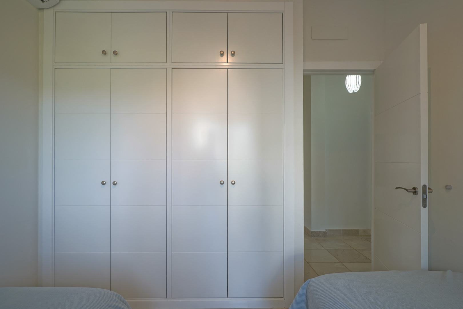 Einbaukleiderschrank in Weiß mit silbernen Griffen und Oberschränken, neben der Zimmertür mit Blick auf den gefliesten Flur.