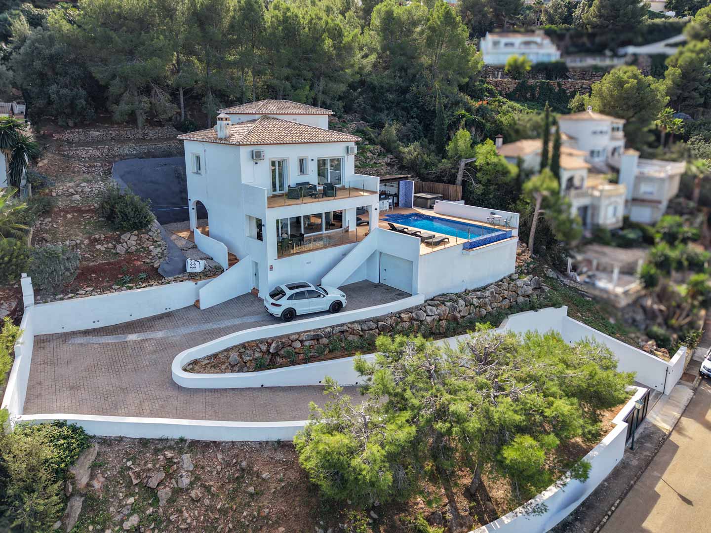 Erhöhte Ansicht der Villa mit weißer Fassade, privatem Pool, Glasgeländer-Terrassen und gepflasterter Auffahrt in der bewaldeten Hügellandschaft von La Sella.