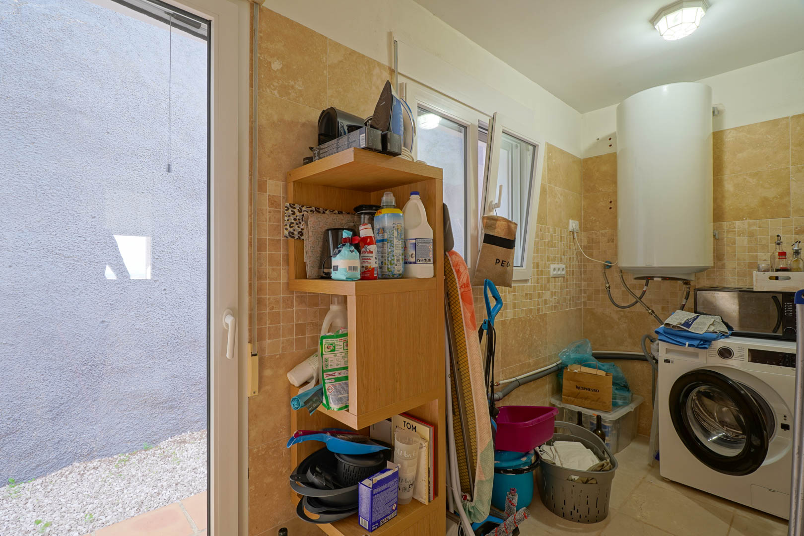 Funktionaler Hauswirtschaftsraum mit cremefarbenen Fliesenwänden, ausgestattet mit Elektroboiler, Waschmaschine, Holzregal und direktem Zugang zum Außenbereich.