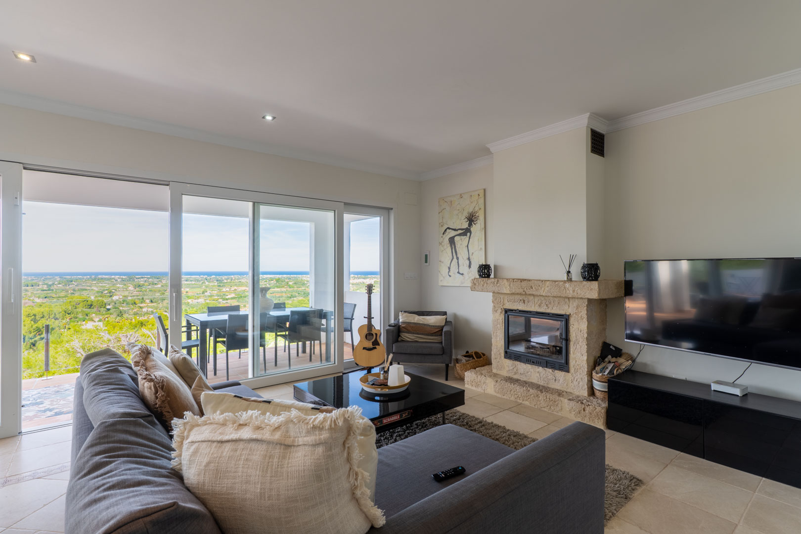 Helles Wohnzimmer mit Natursteinkamin, großen Glasschiebetüren zur Terrasse und Panoramablick auf das Mittelmeer und die umliegende Landschaft.