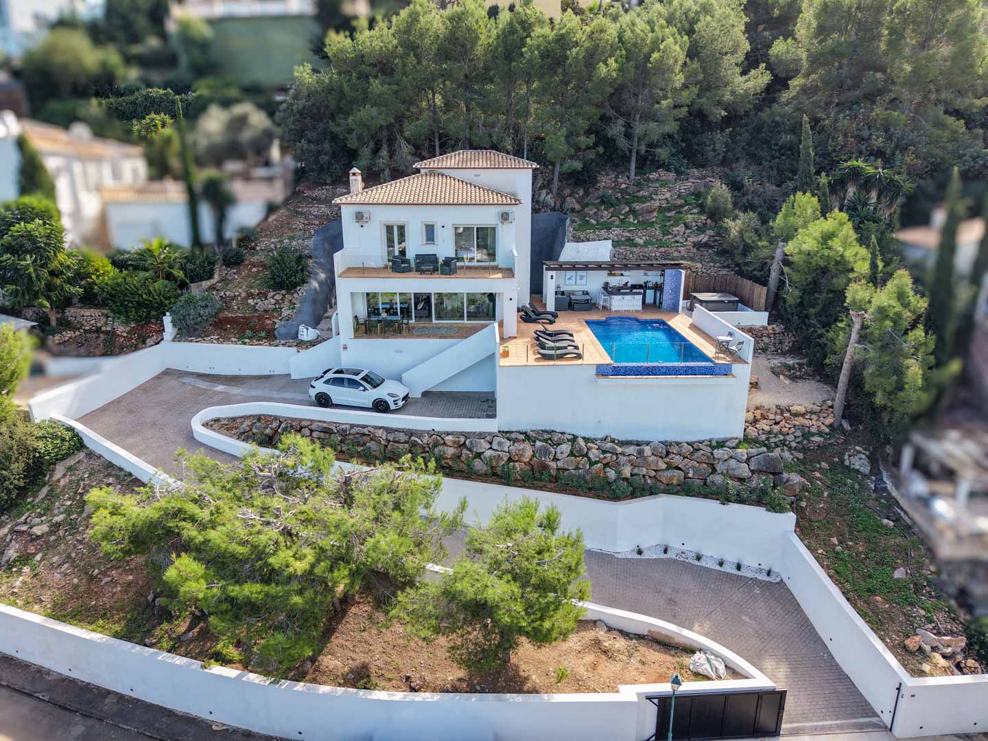 Luftaufnahme der mediterranen Villa mit Infinity-Pool, Außenküche und weitläufigen Terrassen, eingebettet in ein bewaldetes Hanggrundstück in La Sella.