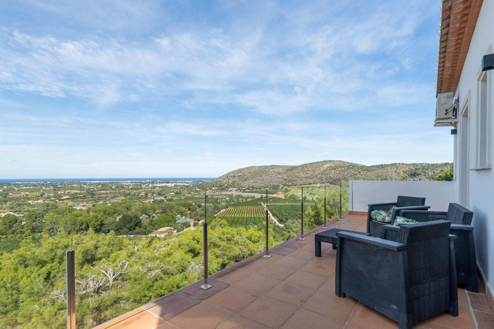 Obere Terrasse mit Glasbalustrade, dunklen Rattanmöbeln und Panoramablick auf das Mittelmeer, den Golfplatz und die umliegende Küstenlandschaft von La Sella.