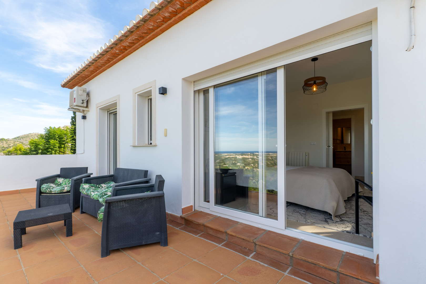 Obere Terrasse mit Terrakotta-Fliesen und Gartenmöbeln, die durch große Glasschiebetüren direkten Zugang zum Schlafzimmer und einen weiten Meerblick bietet.
