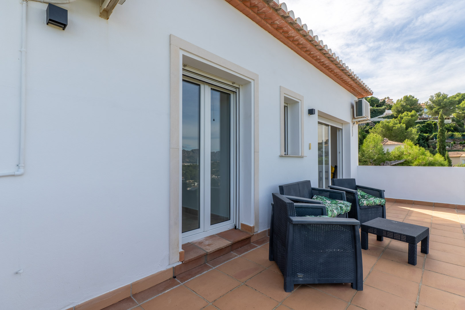 Obere Terrasse mit Terrakottafliesen, dunklen Polyrattan-Sitzmöbeln und direktem Zugang zu den Schlafzimmern durch großzügige Glastüren.