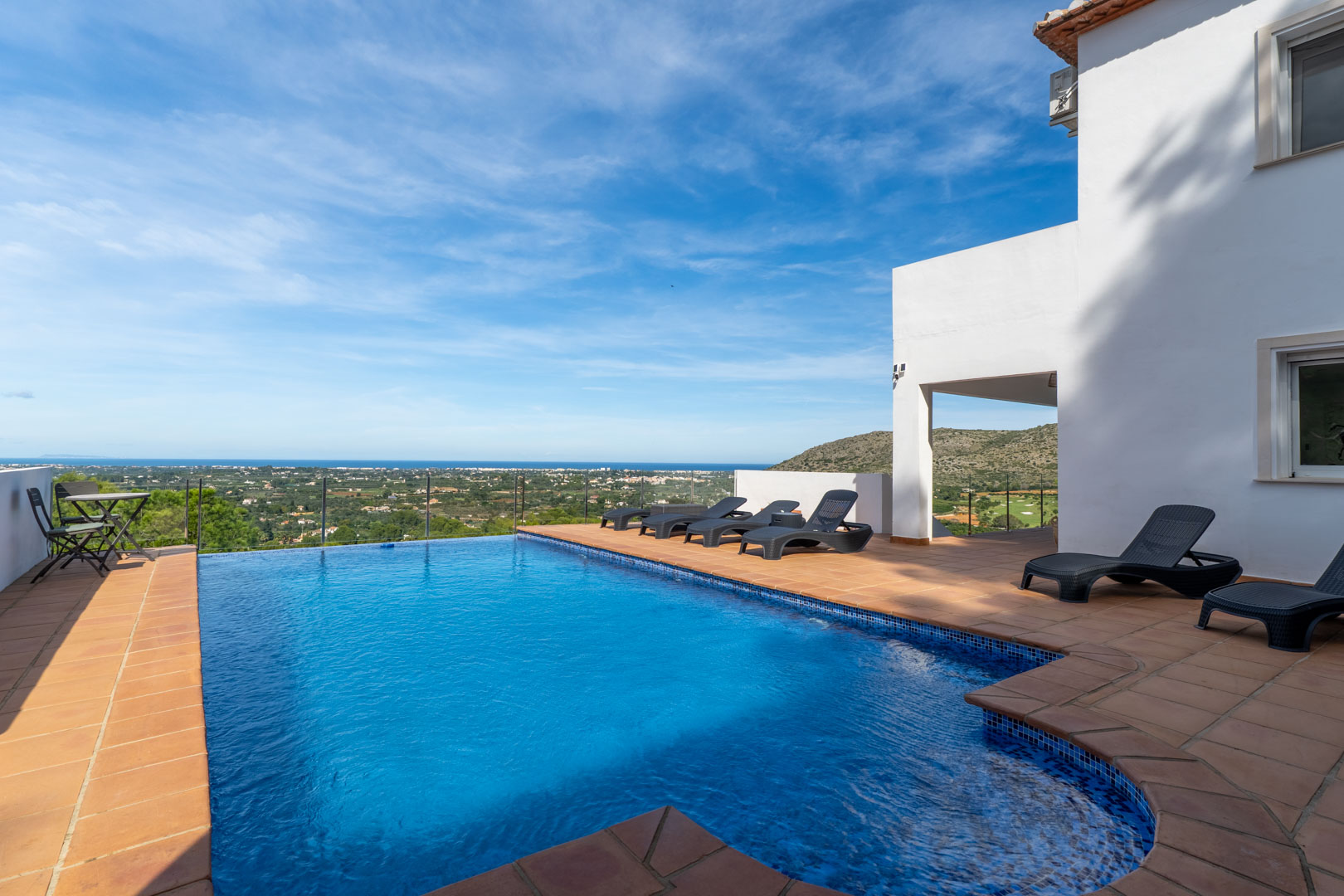 Privater Pool mit Terrakotta-Terrasse, schwarzen Sonnenliegen und Glasgeländer mit Panoramablick auf das Mittelmeer, das Tal und den Golfplatz.