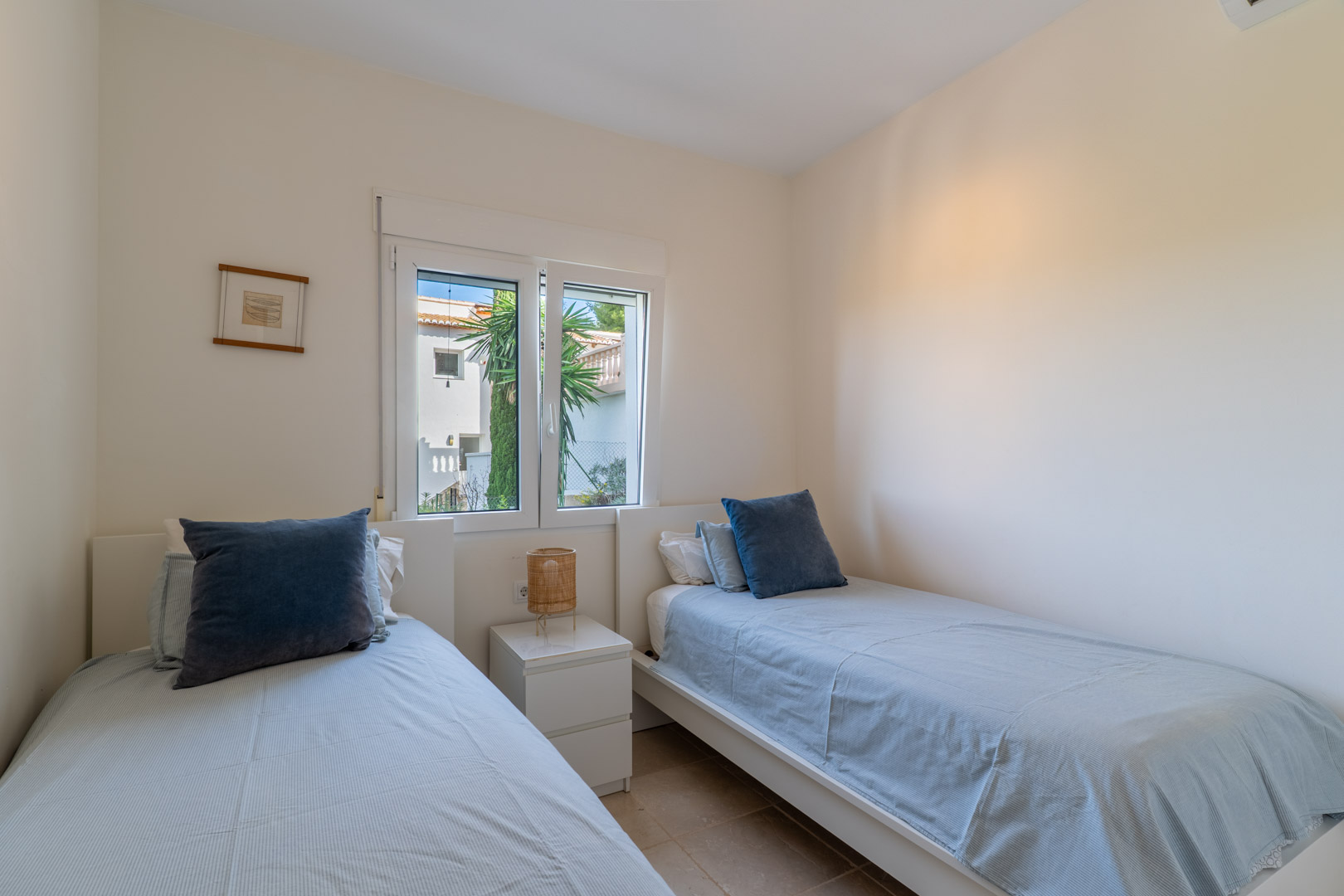 Schlafzimmer mit zwei Einzelbetten, weißem Nachttisch und Fliesenboden. Das Fenster sorgt für Tageslicht und bietet Ausblick auf die Umgebung.