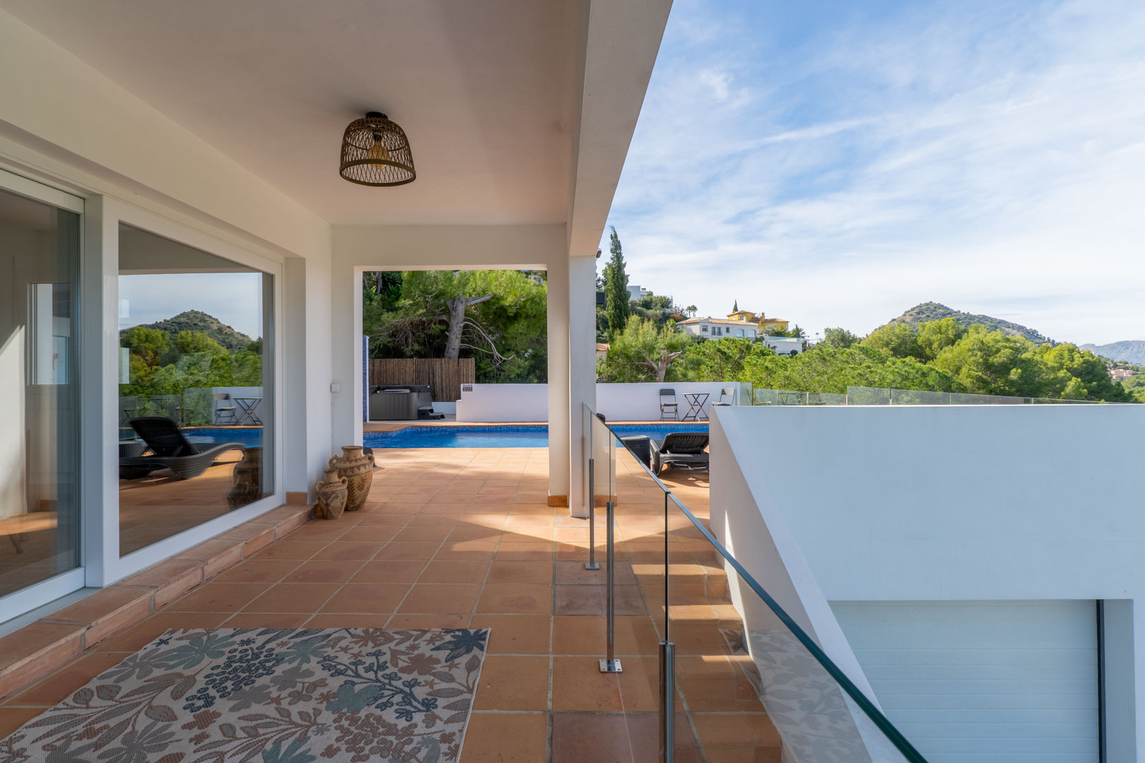 Überdachte Terrasse mit Terrakottaboden, Glasgeländer und Blick auf den privaten Pool. Dekorative Tonkrüge und eine Rattanlampe unterstreichen das mediterrane Design.