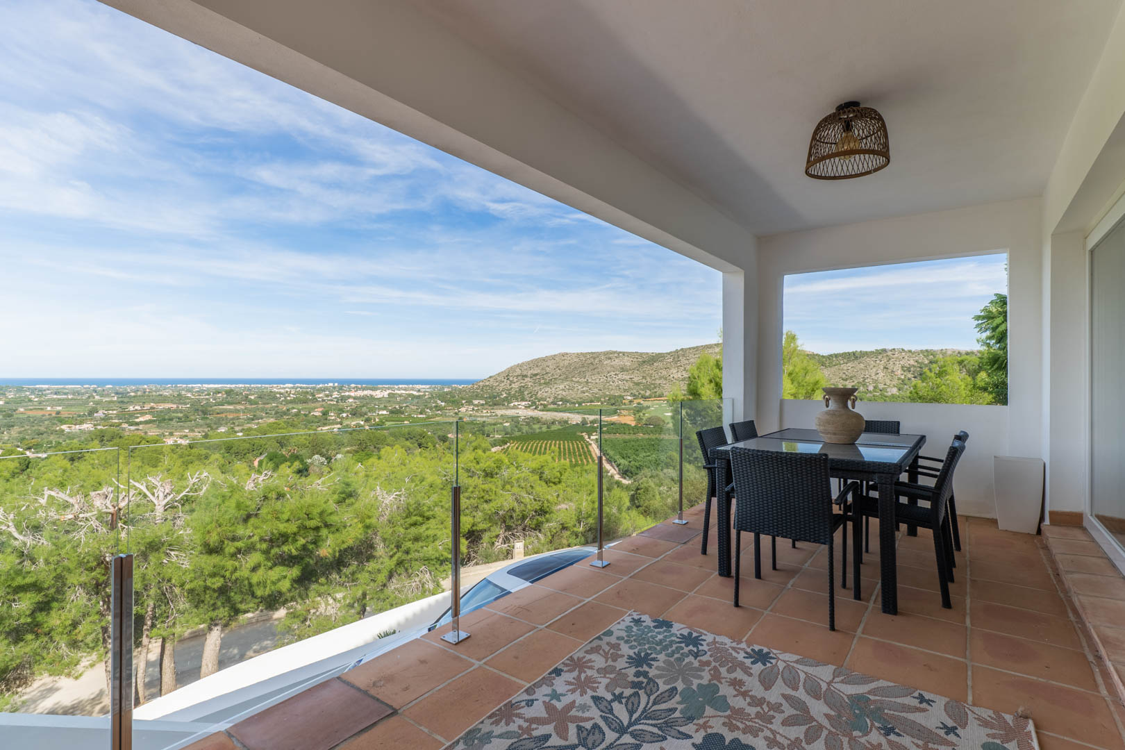 Überdachte Terrasse mit Terrakottafliesen, Essgruppe und Glasgeländer mit Panoramablick auf das Mittelmeer und die umliegende Hügellandschaft.
