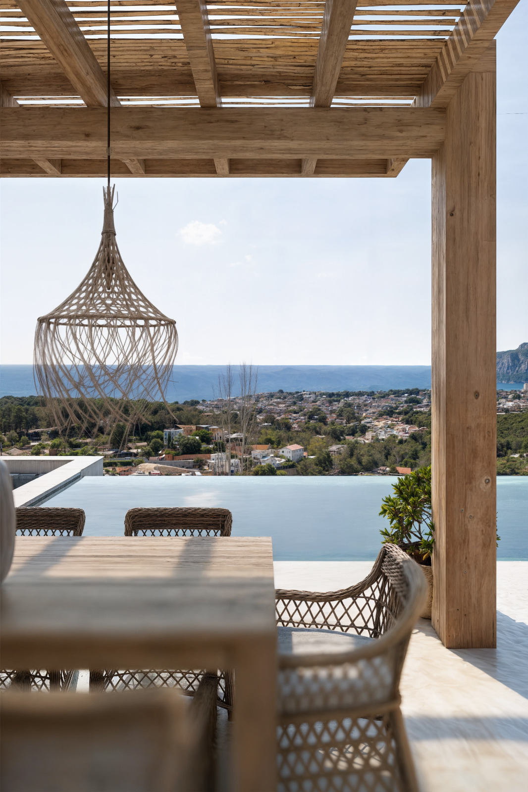 Außen-Terrasse mit Holzpergola, Essbereich aus Korbgeflecht und einer Hängeleuchte aus Naturfasern mit Blick auf den Infinity-Pool und das Meer.
