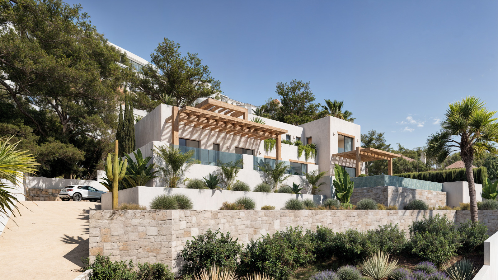 Außenansicht der Villa im Ibiza-Stil mit weißen Fassaden, Holzpergolen und Natursteinmauern, eingebettet in einen mediterranen Garten mit Palmen.