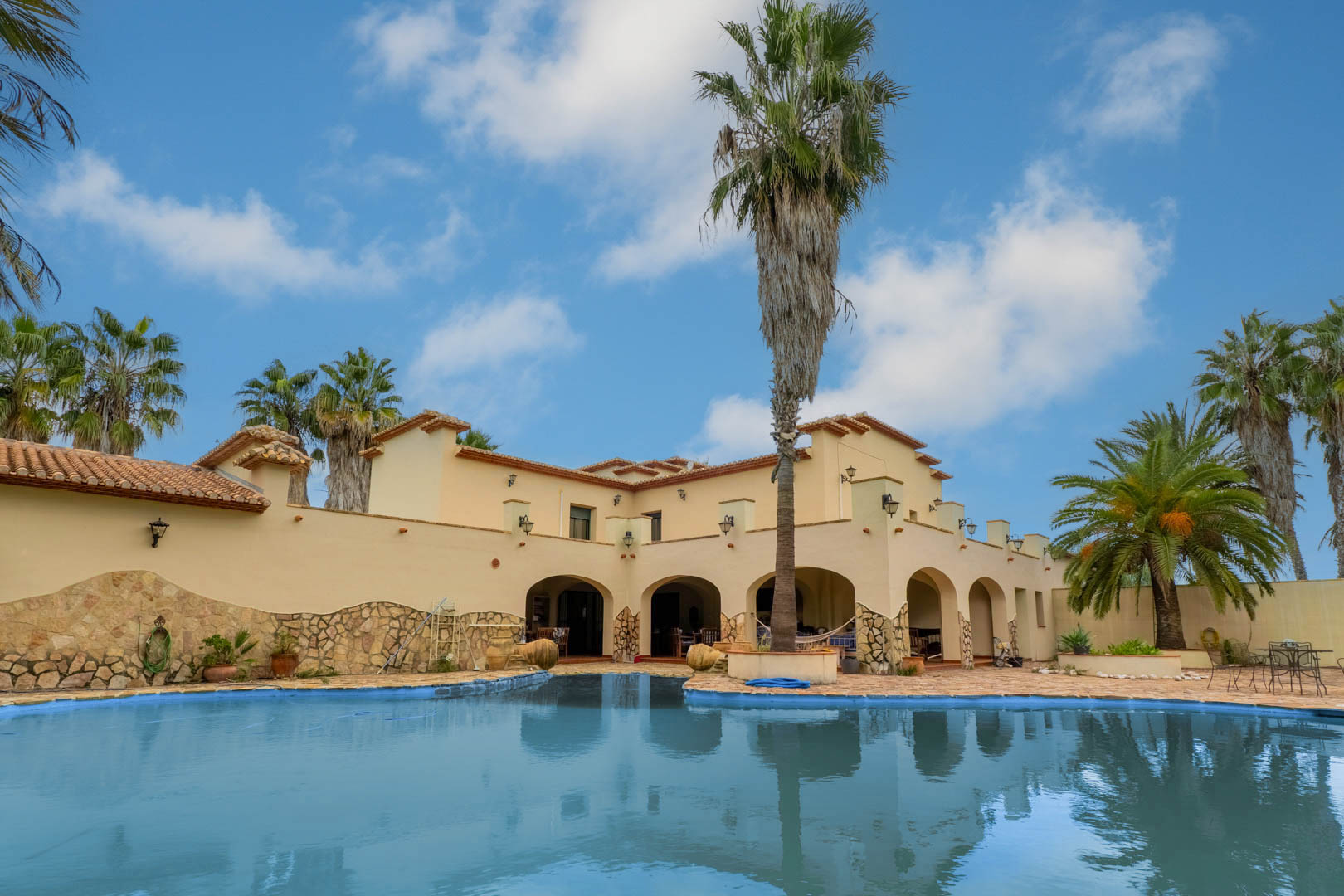 Außenansicht des geschwungenen Swimmingpools mit Steinterrasse, der mediterranen Fassade der Villa und den markanten Rundbogen-Veranden unter Palmen.