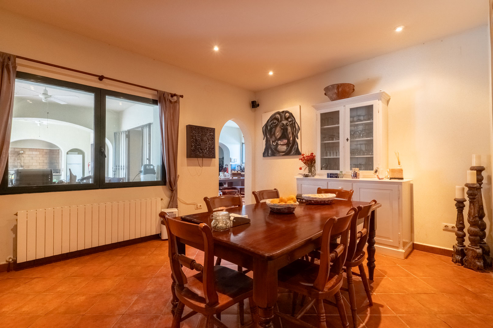 Essbereich mit Terrakottaboden, massivem Holztisch und weißem Sideboard. Ein großes Fenster blickt auf die Veranda, ergänzt durch dezente Deckenstrahler.