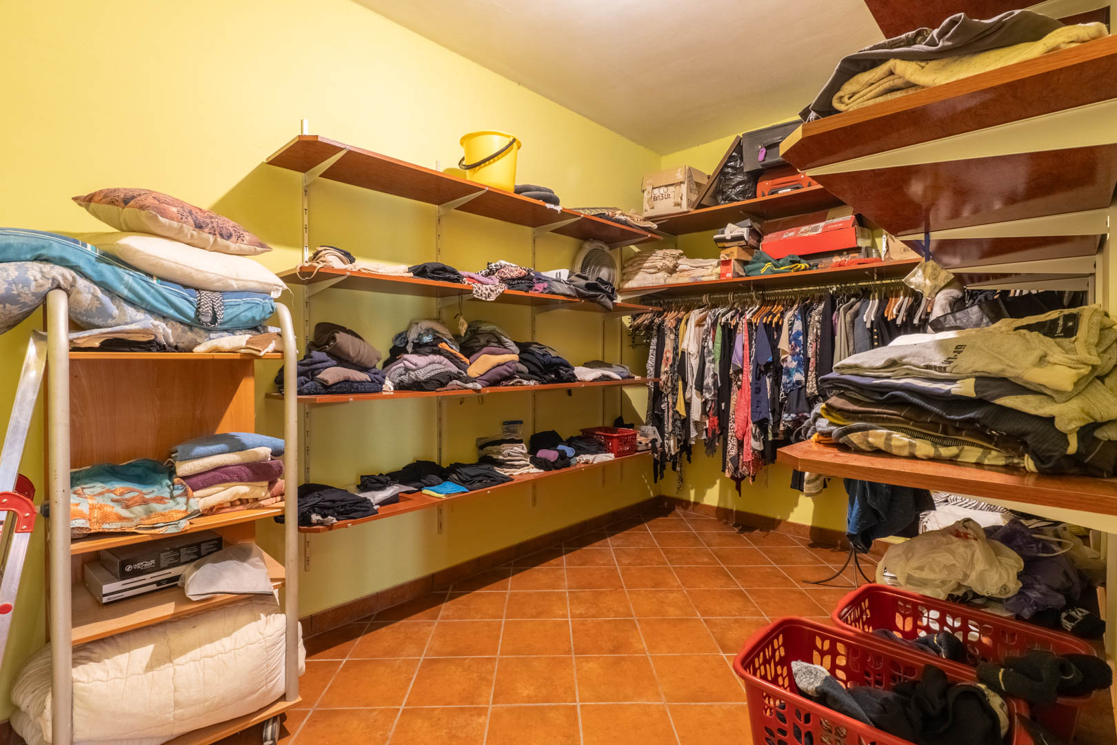 Funktionaler begehbarer Kleiderschrank mit umlaufenden Holzregalen, Kleiderstangen und rustikalem Terrakottaboden für großzügigen Stauraum.