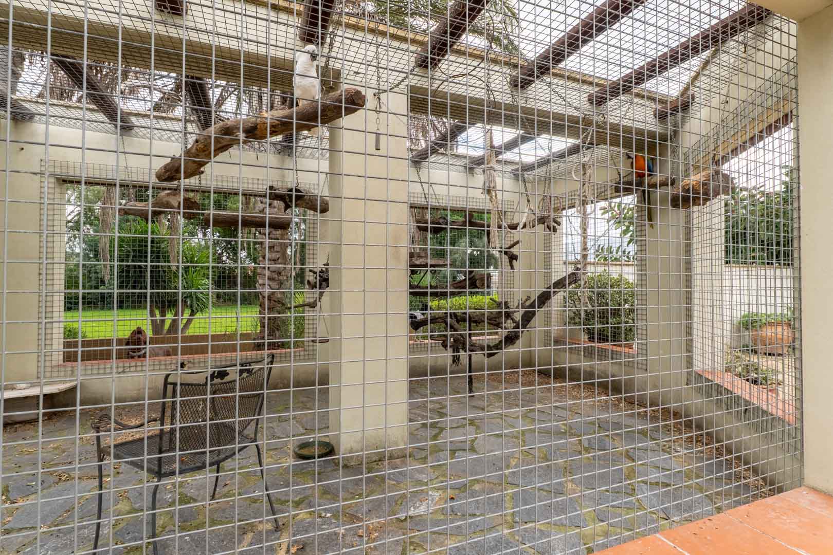 Große Außenvoliere mit Metallgitter, Schieferboden und Naturholzstangen, eingebettet in die gepflegte Gartenanlage des Anwesens.