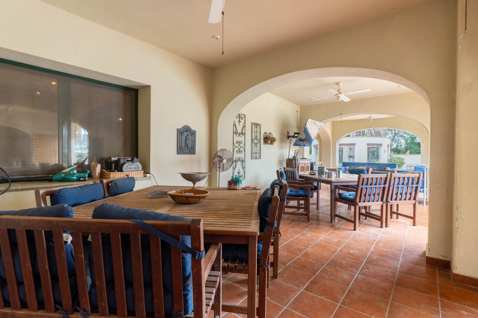 Großzügige überdachte Terrasse mit mediterranen Rundbögen, Terrakottaboden und Holzmöbeln. Ausgestattet mit Deckenventilatoren und direktem Blick in den weitläufigen Garten.