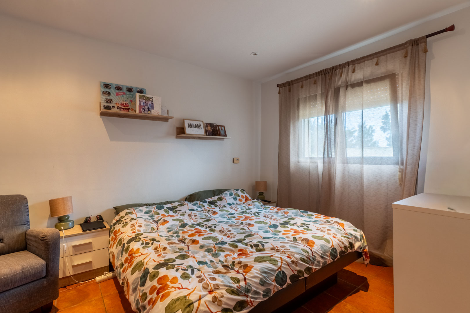 Helles Schlafzimmer mit Terrakotta-Fliesenboden, weißen Wänden und Holzregalen. Ein Fenster mit lichtdurchlässigen Vorhängen und Rollladen sorgt für natürliches Licht.