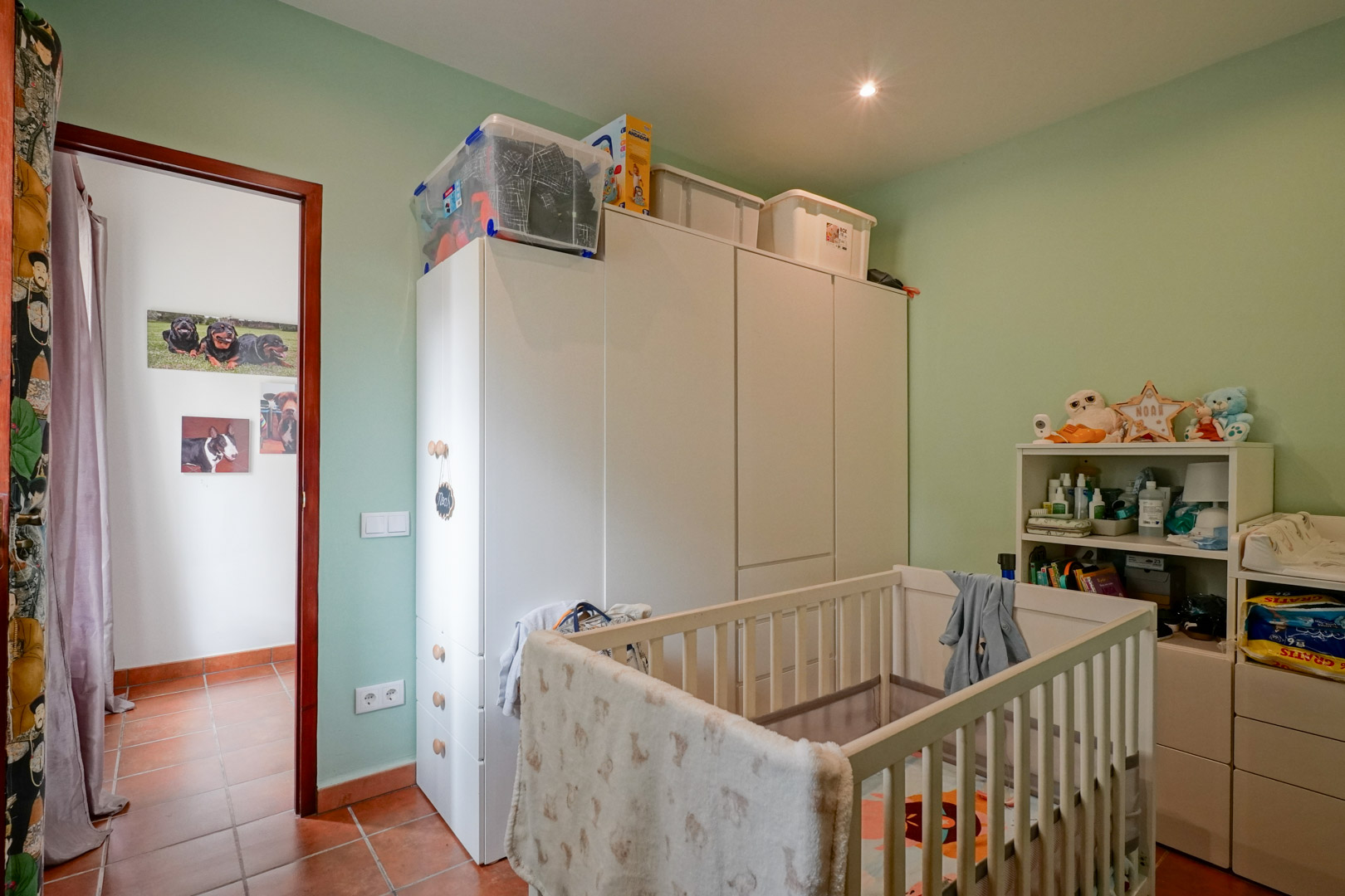 Kinderzimmer mit weißem Holzgitterbett, passenden Kleiderschränken und hellgrünen Wänden. Der Raum verfügt über traditionelle Terrakotta-Fliesen und Deckenstrahler.