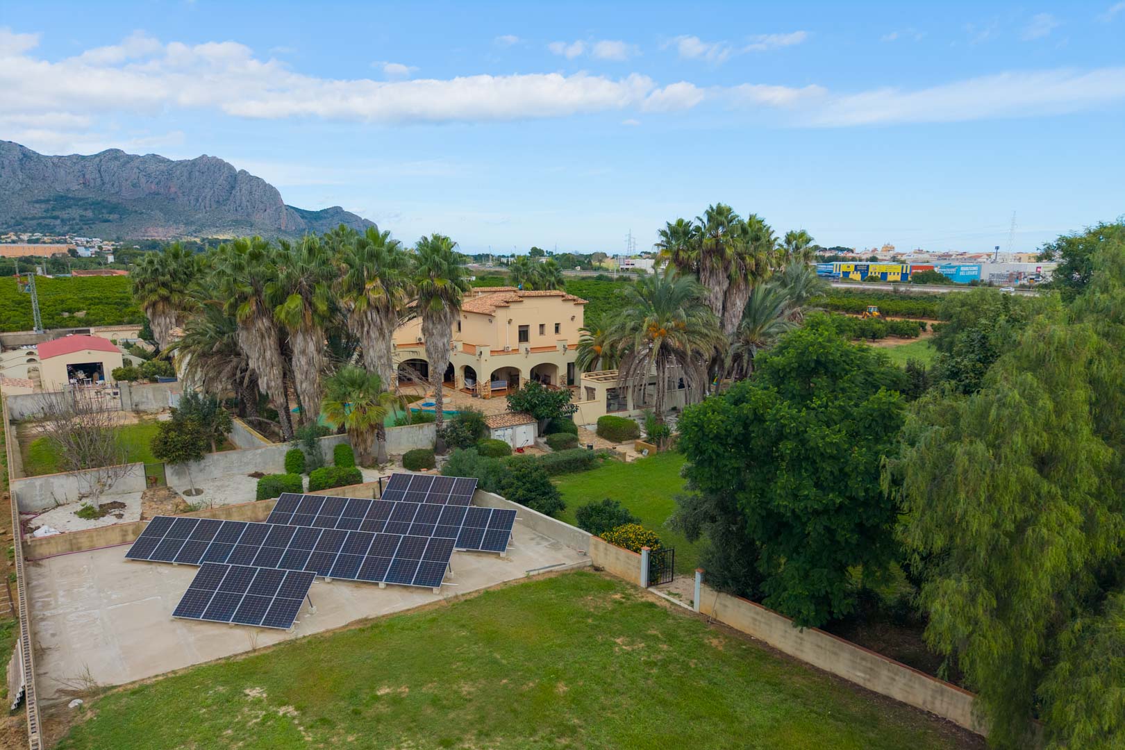 Luftaufnahme des Anwesens mit Photovoltaikanlage, weitläufigen Rasenflächen, Palmen und der mediterranen Villa vor einer Bergkulisse.