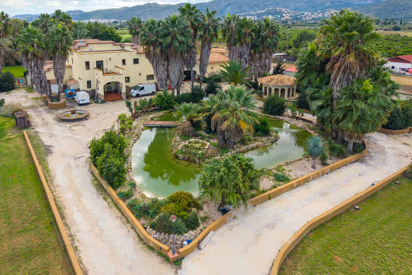 Luftaufnahme des Haupteingangs mit einem großen Zierteich, einer Holzbrücke und hohen Palmen vor der mediterranen Villa.