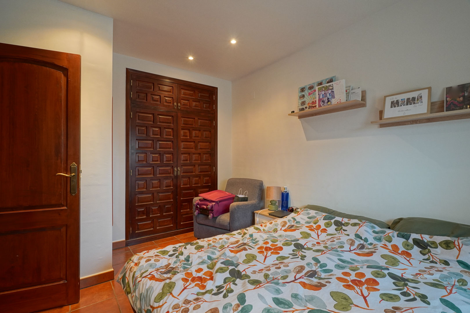 Schlafzimmer mit einem Einbauschrank aus dunklem Massivholz, Terrakottaboden und Deckenspots. Schlichte Wandregale ergänzen das traditionelle Design.