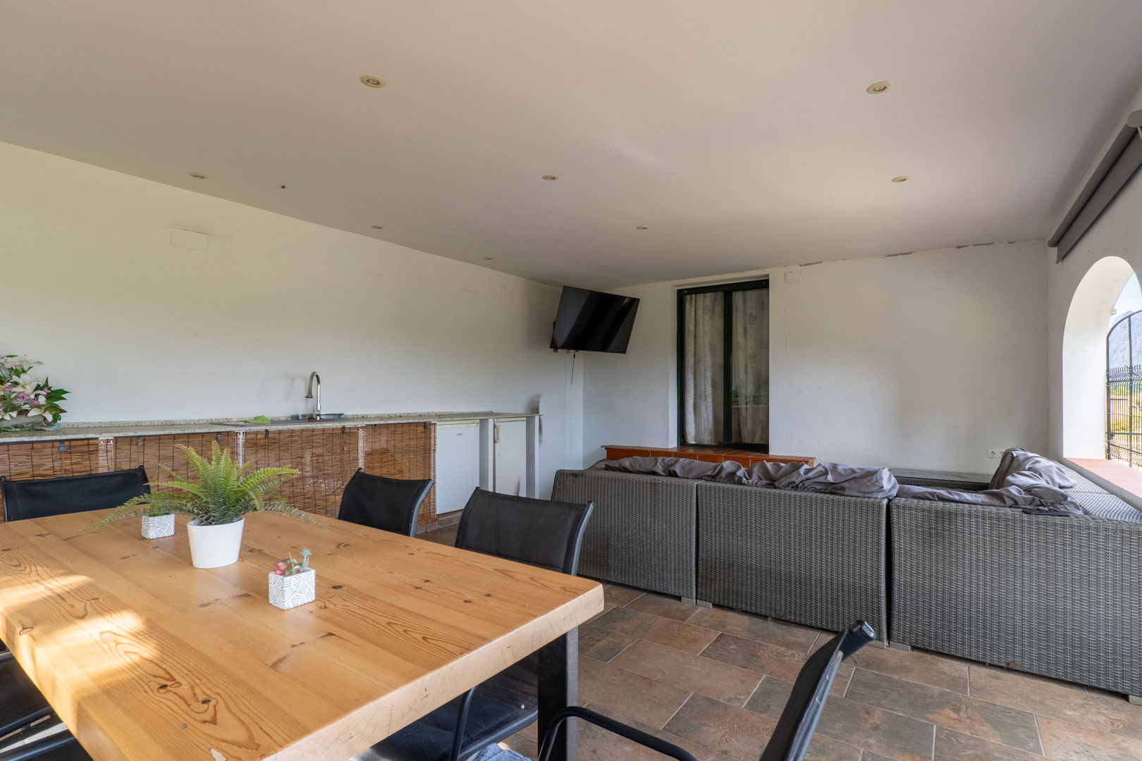 Überdachte Terrasse mit grauer Rattan-Sitzgruppe, massivem Holzesstisch und Außenküche. Der Bereich ist mit Deckeneinbaustrahlern und rustikalen Bodenfliesen ausgestattet.