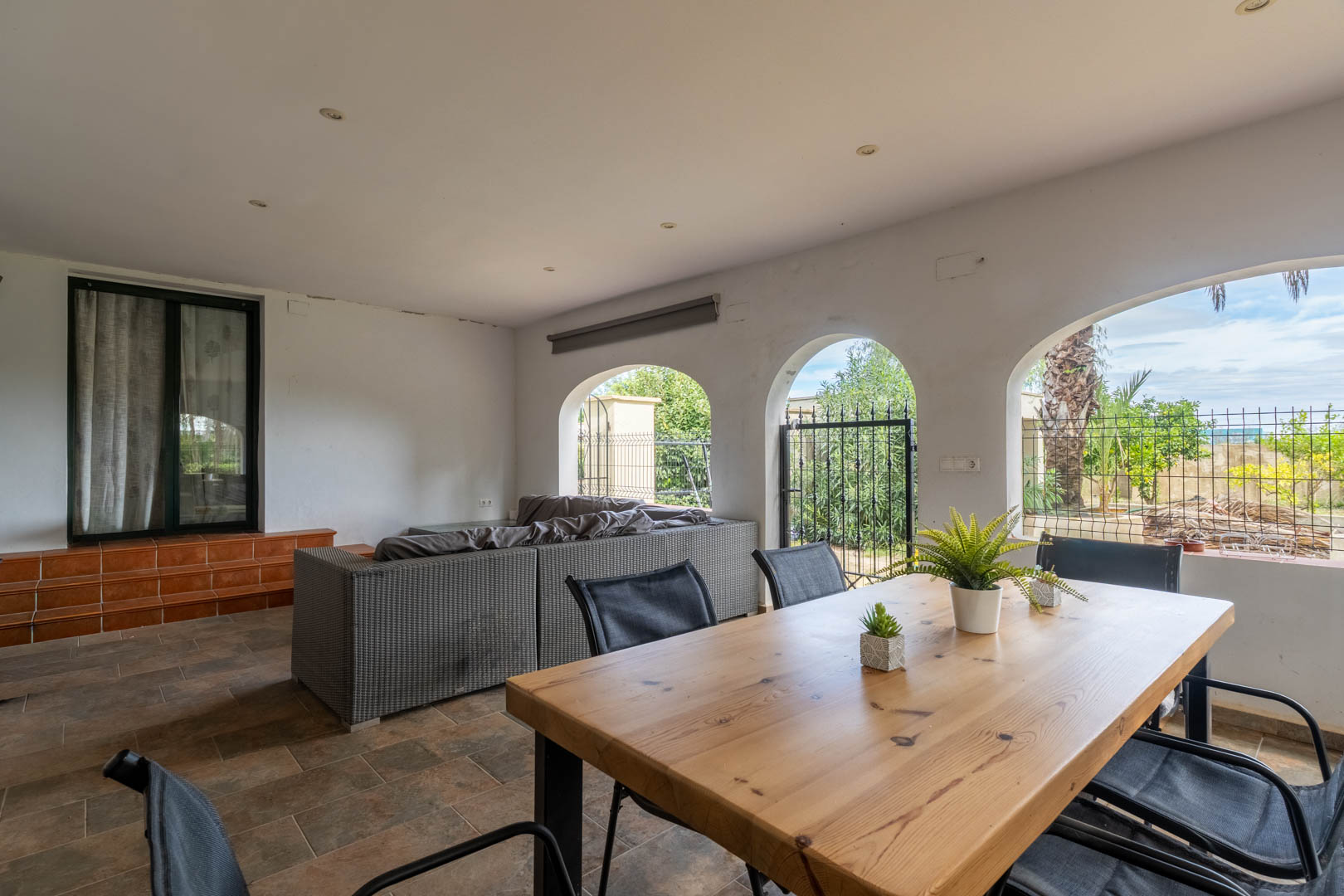 Überdachte Terrasse mit mediterranen Rundbögen, Massivholztisch, Rattan-Sofas und Gartenblick. Rustikaler Fliesenboden und Deckeneinbaustrahler.