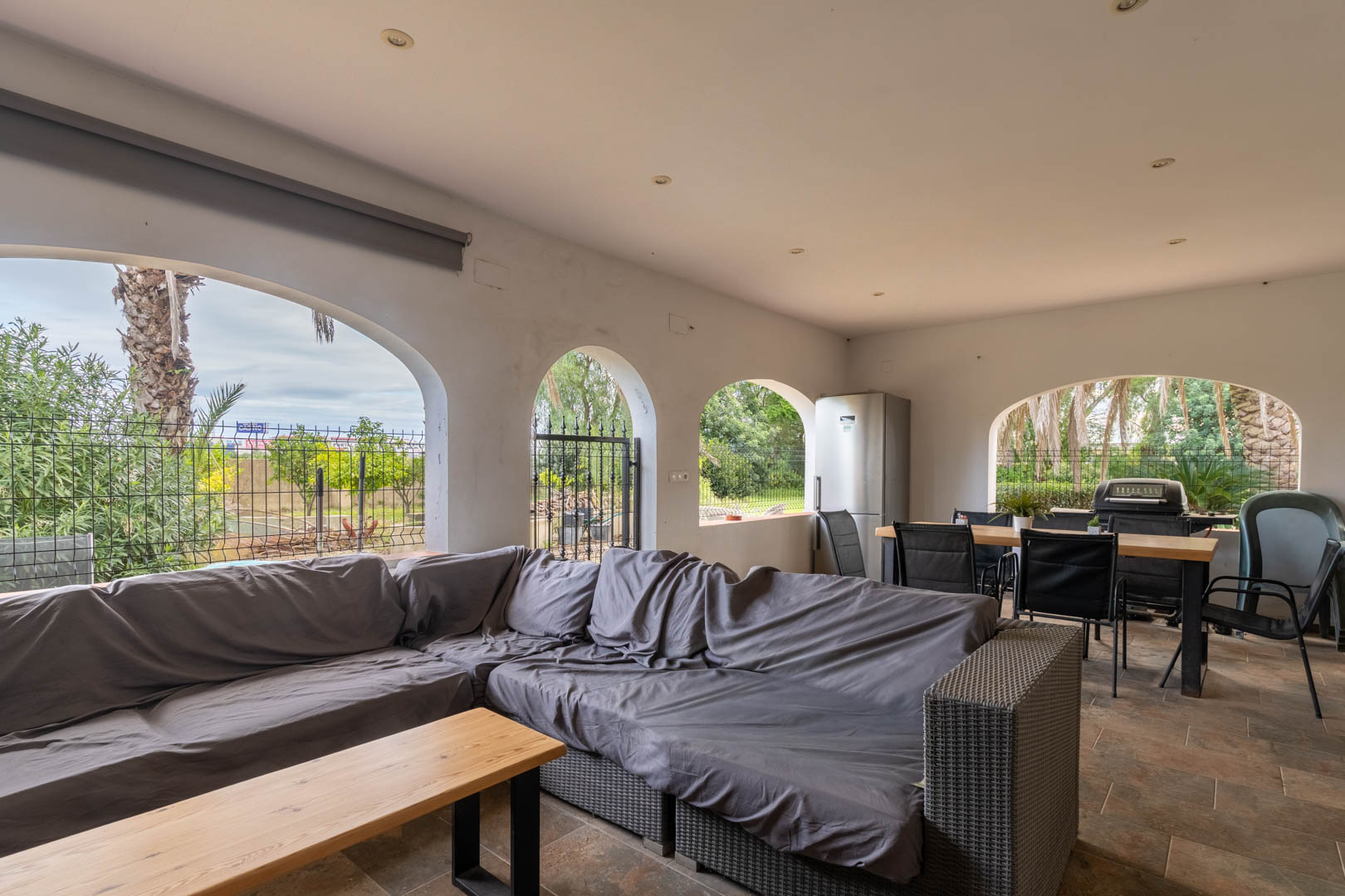 Überdachte Terrasse mit Rundbögen, grauer Lounge-Ecke und Außen-Essbereich mit Grill und Kühlschrank. Fliesenboden und Deckeneinbaustrahler mit Gartenblick.