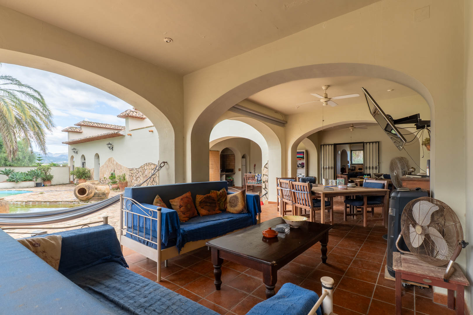 Überdachte Terrasse mit Rundbögen, Terrakotta-Fliesen und Loungebereich. Der Raum bietet Blick auf den Pool, die Hängematte und den mediterranen Garten mit Palmen.
