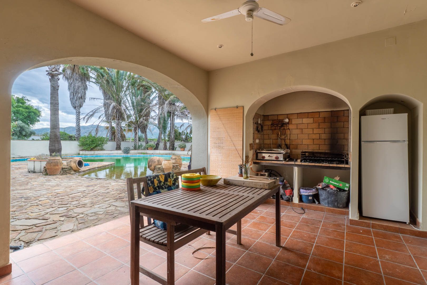 Überdachte Veranda mit gemauertem Grill, Esstisch aus Holz und Blick durch einen Rundbogen auf den Pool und die weitläufigen Gartenanlagen.