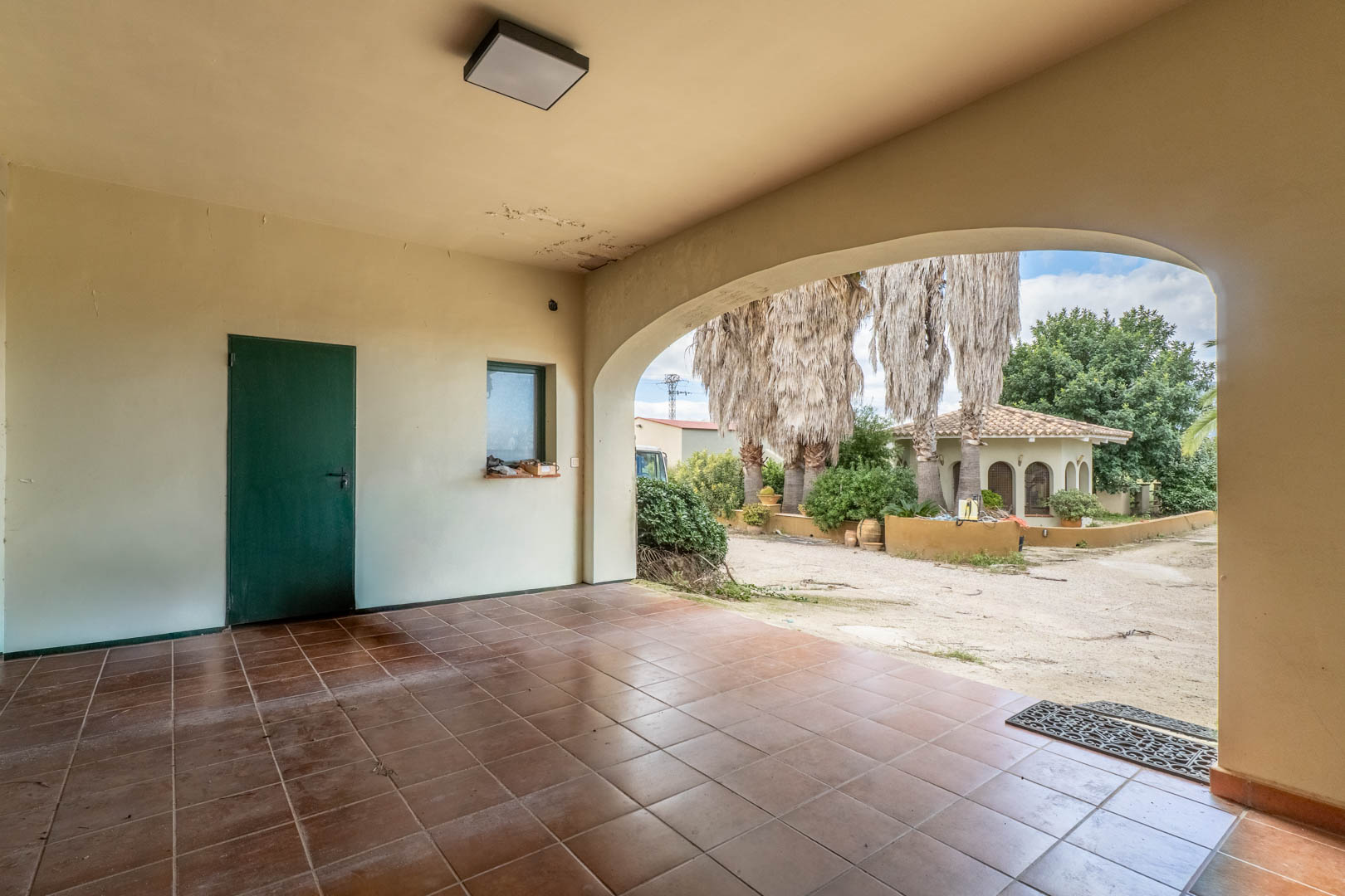 Überdachte Veranda mit Terrakotta-Fliesen und Rundbogen, der den Blick auf die mediterranen Gärten und das separate Gästehaus freigibt.
