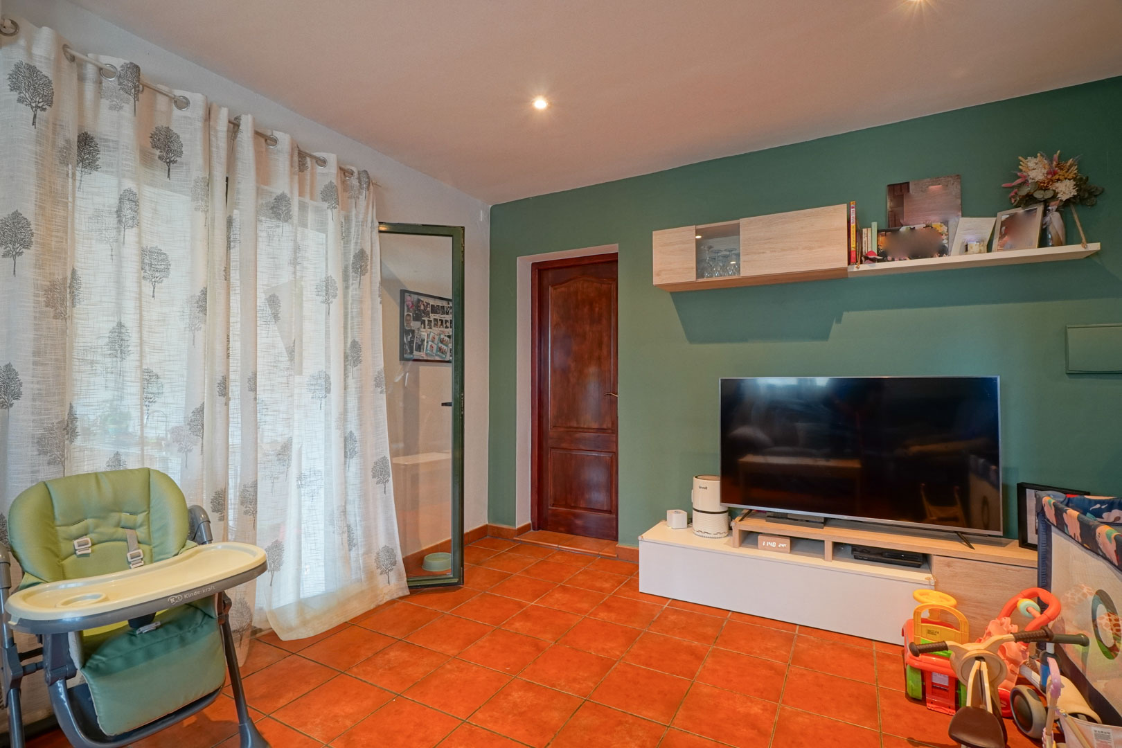 Wohnbereich mit salbeigrüner Akzentwand, Terrakotta-Fliesenboden und modernem TV-Board. Eine Massivholztür und Einbaustrahler prägen den funktionalen Raum.