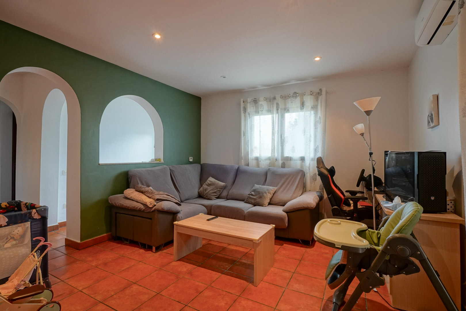 Wohnbereich mit Terrakottafliesen, grüner Akzentwand und Rundbogen-Durchbrüchen. Ausgestattet mit Ecksofa, Arbeitsbereich und Split-Klimaanlage.