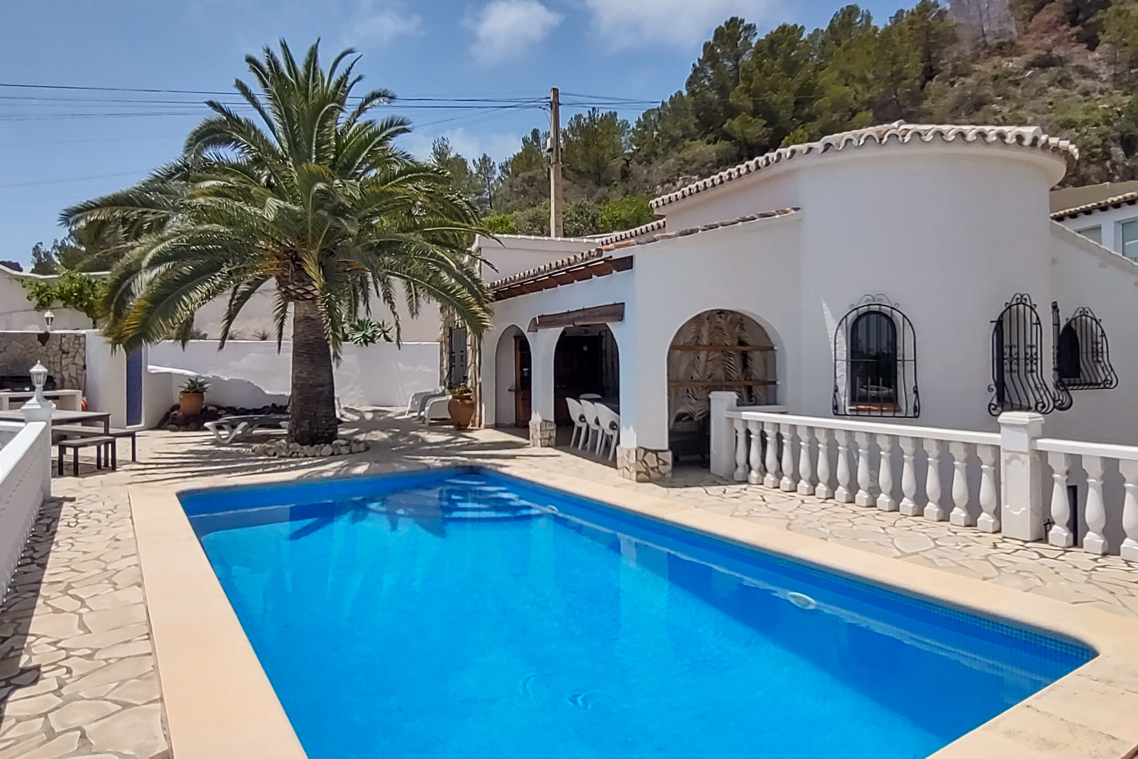 Außenansicht des rechteckigen Pools und der Natursteinterrasse mit der weißen Villa, traditionellen Rundbögen, schmiedeeisernen Fenstergittern und einer großen Palme.