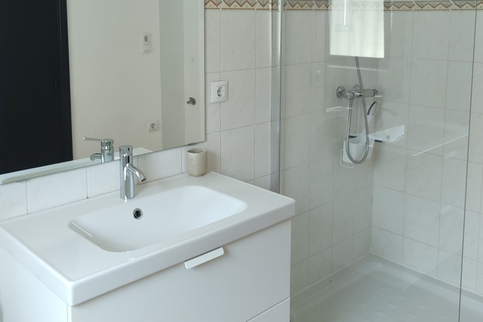 Funktionales Badezimmer mit weißem Waschtisch, verchromten Armaturen und einer begehbaren Dusche mit Glaswand. Weiße Fliesen mit dekorativer Bordüre an den Wänden.