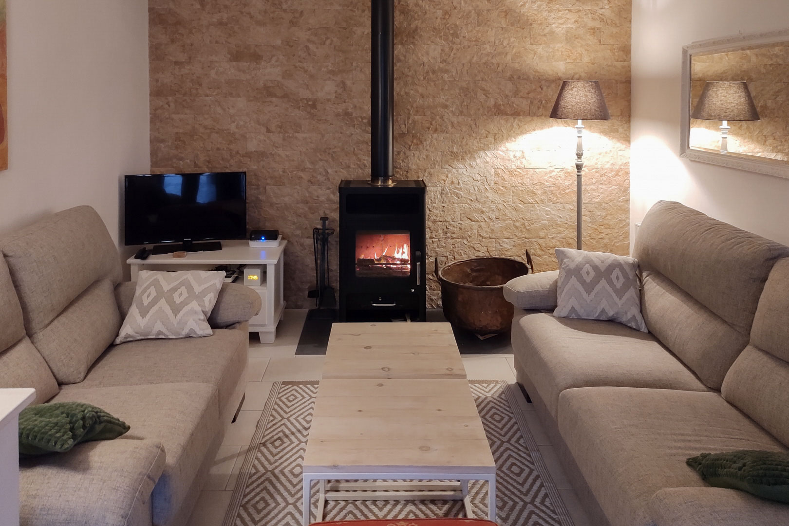 Gemütlicher Wohnbereich mit Kaminofen vor einer Natursteinwand, ausgestattet mit neutralen Sofas und einem Couchtisch aus hellem Holz.