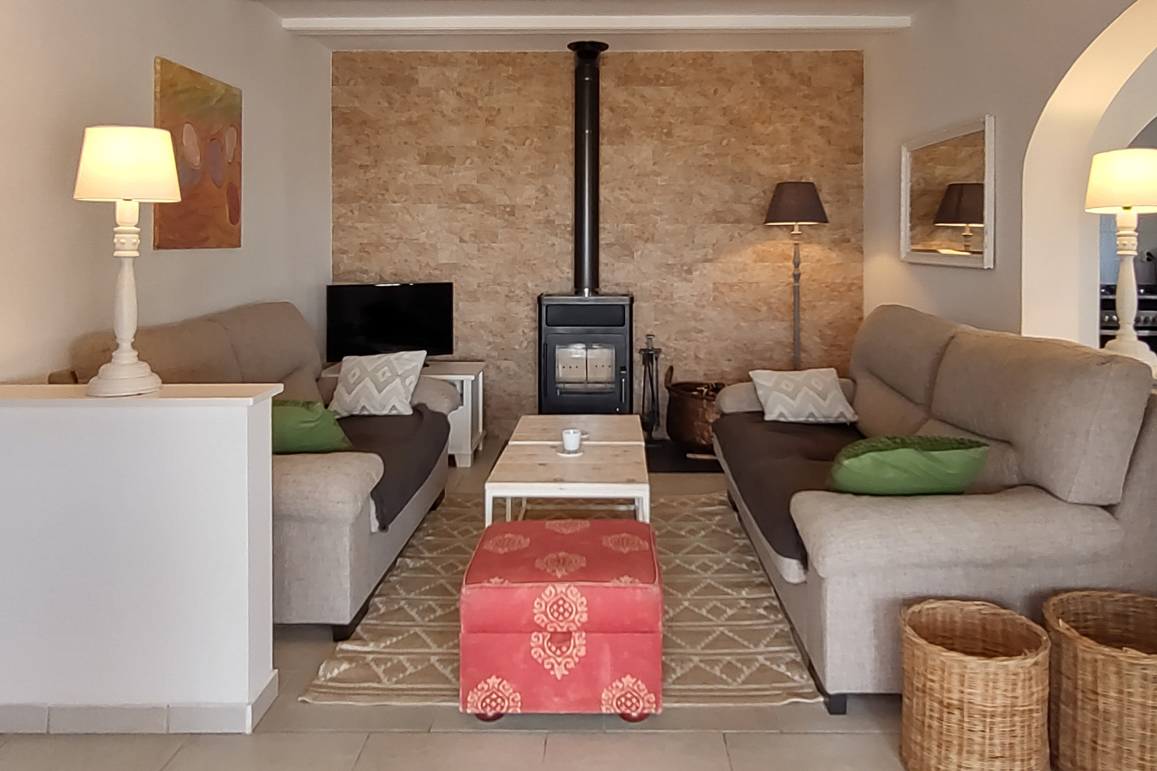 Gemütliches Wohnzimmer mit Holzofen vor einer Natursteinwand, gegenüberliegenden Sofas, Holzcouchtisch und einem roten dekorativen Polsterhocker.