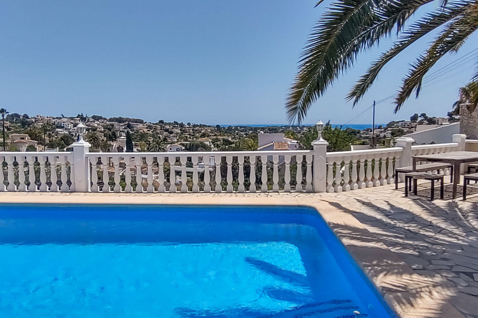 Poolterrasse mit weißer Balustrade und Panoramablick auf das Meer und das Tal, ausgestattet mit Natursteinpflaster und einer hölzernen Sitzgruppe.