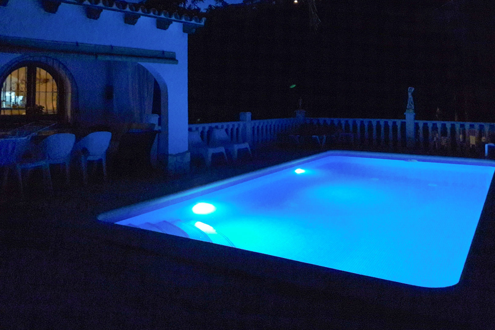 Rechteckiger Swimmingpool mit blauer LED-Unterwasserbeleuchtung, angrenzend an eine überdachte Bogenterrasse und eine klassische weiße Balustrade.