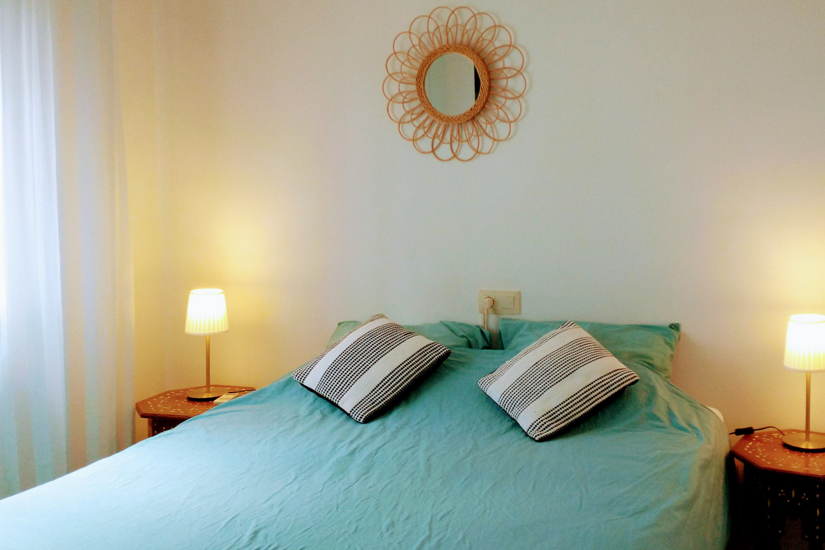 Schlafzimmer mit Doppelbett, türkisfarbener Bettwäsche, hexagonalen Holznachttischen und einem dekorativen Rattanspiegel an der weißen Wand.