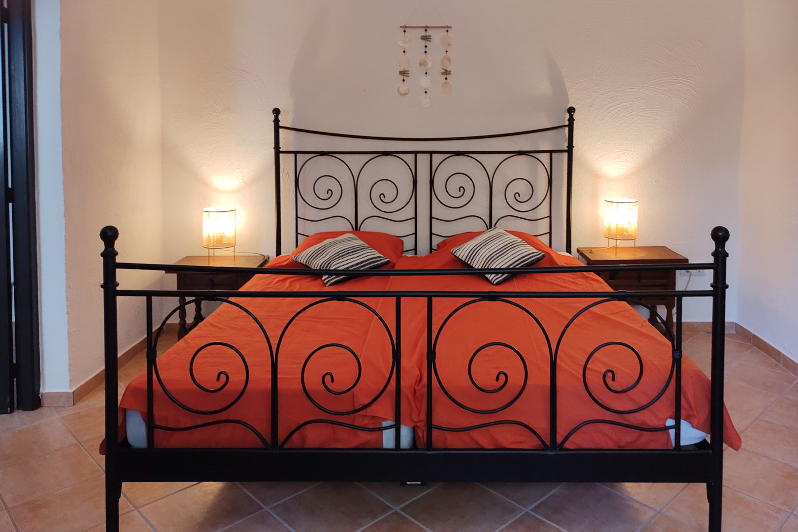 Schlafzimmer mit schwarzem Schmiedeeisenbett, Holznachtischen und Fliesenboden. Warme Nachttischlampen setzen die weiß strukturierten Wände dezent in Szene.