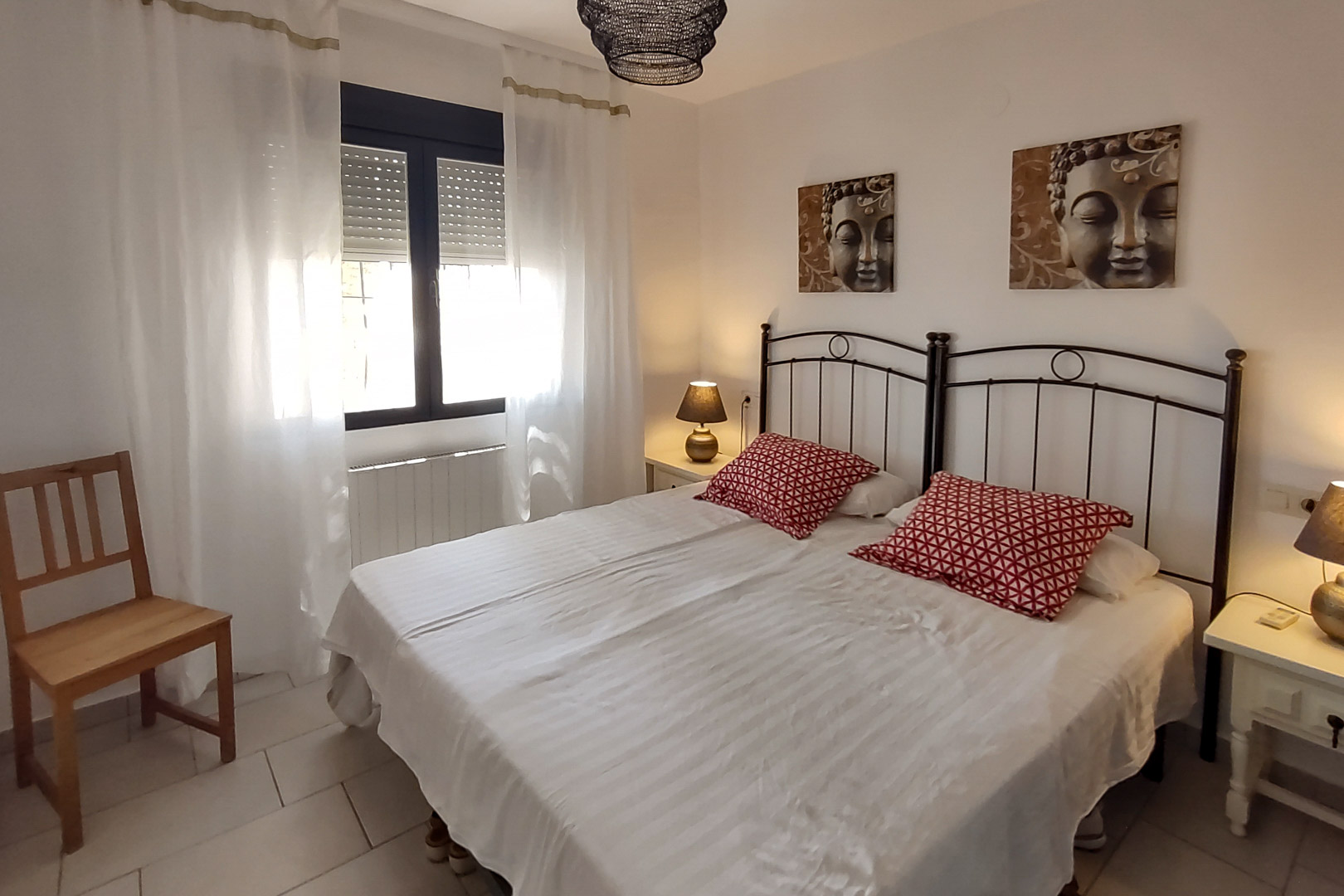 Schlafzimmer mit zwei Einzelbetten, schwarzen schmiedeeisernen Kopfteilen, Buddha-Bildern und Fenster mit Rollläden und weißen Vorhängen.