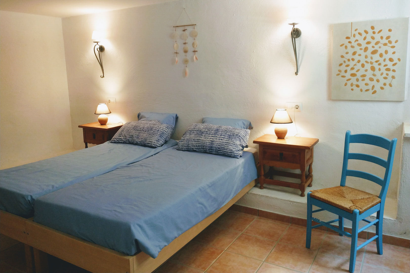 Schlafzimmer mit zwei Einzelbetten, Terrakottaboden und weißen Wänden. Ausgestattet mit Nachttischen aus Holz, Wandleuchten und einem blauen rustikalen Stuhl.