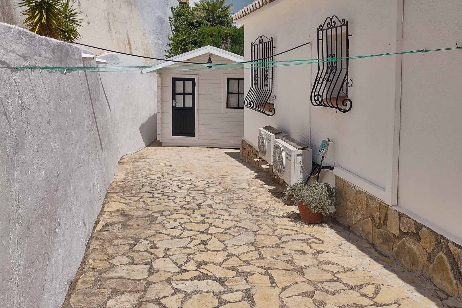 Seitlicher Weg mit Natursteinpflasterung, der zu einem hölzernen Gartenhaus führt, mit Außenklimageräten und traditionellen schmiedeeisernen Fenstergittern.
