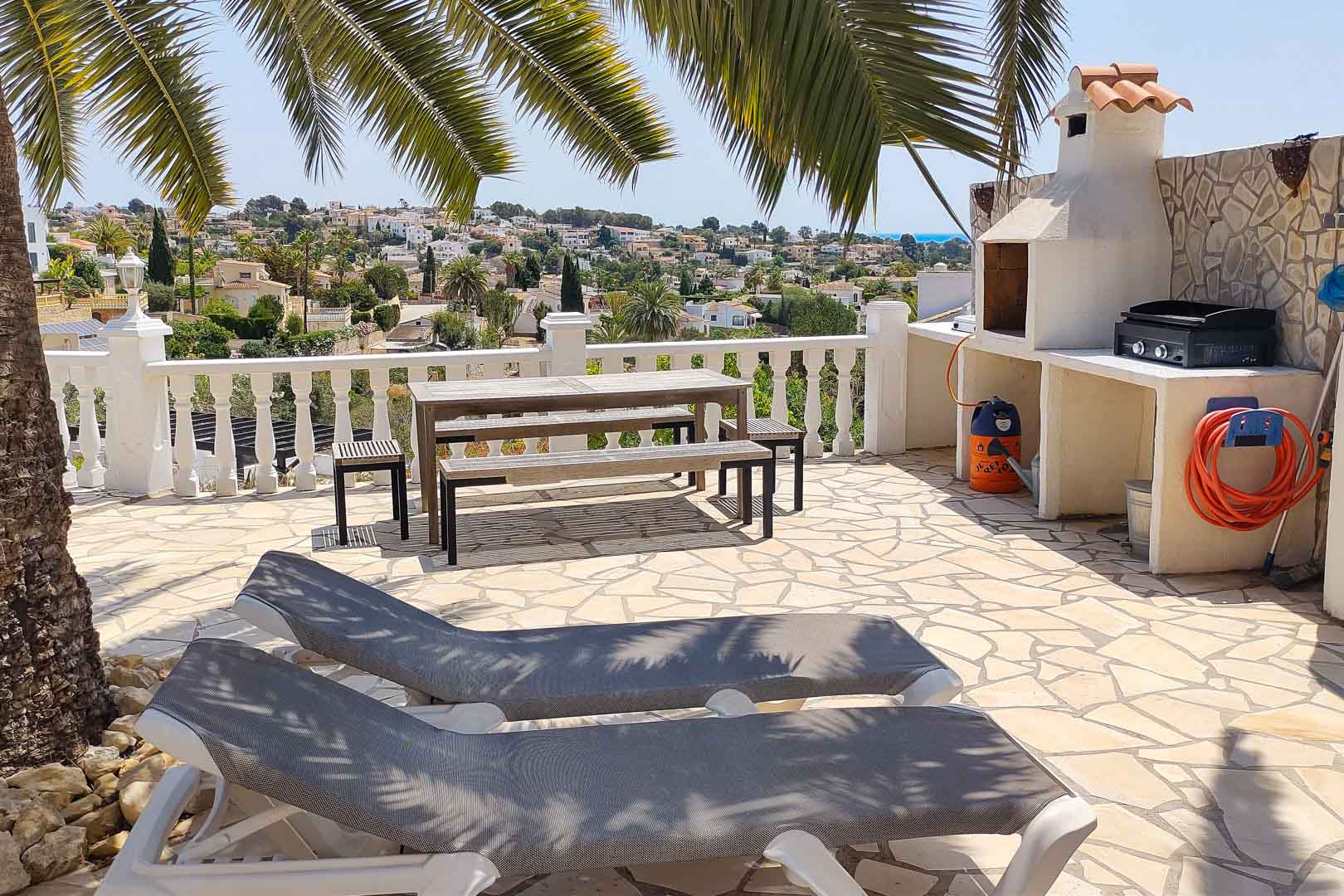 Steinpflaster-Terrasse mit gemauertem Grill, Picknicktisch aus Holz und Sonnenliegen, beschattet von Palmen mit Blick über das Tal bis zum Meer.