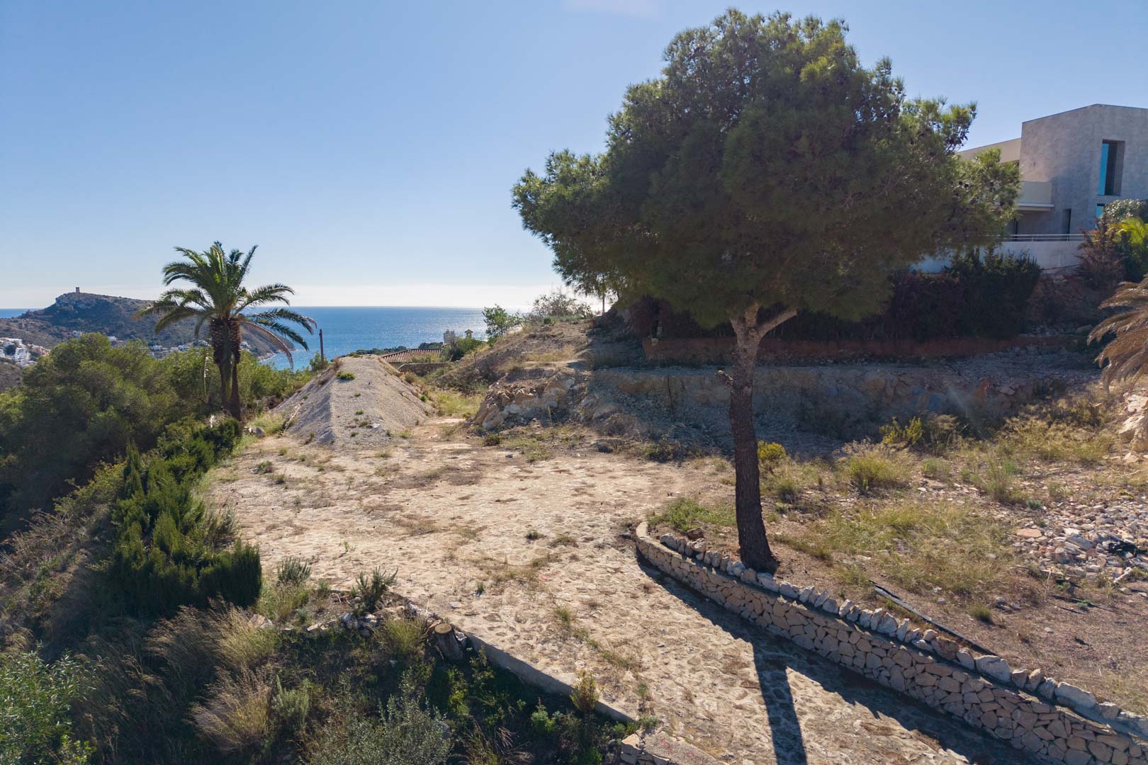 Außenansicht des Grundstücks mit Natursteinweg, Pinien, Palmen und Panoramablick auf das Mittelmeer in Moraira.