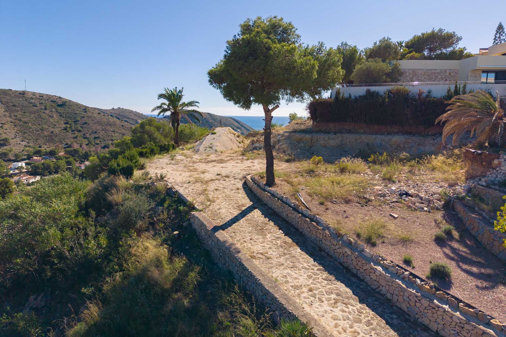 Panoramablick vom Grundstück mit gepflastertem Weg und Trockensteinmauern, umgeben von mediterraner Flora und freiem Meerblick.