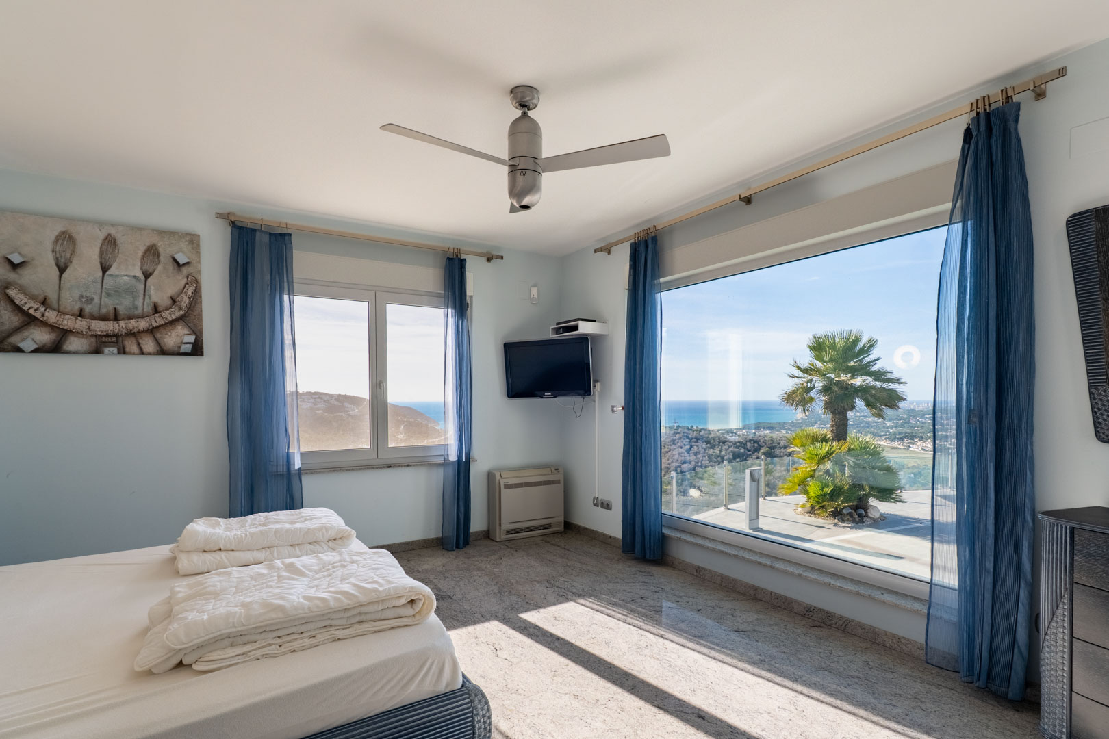 Helles Schlafzimmer mit Granitböden, blauen Vorhängen und Meerblick durch große Fenster.
