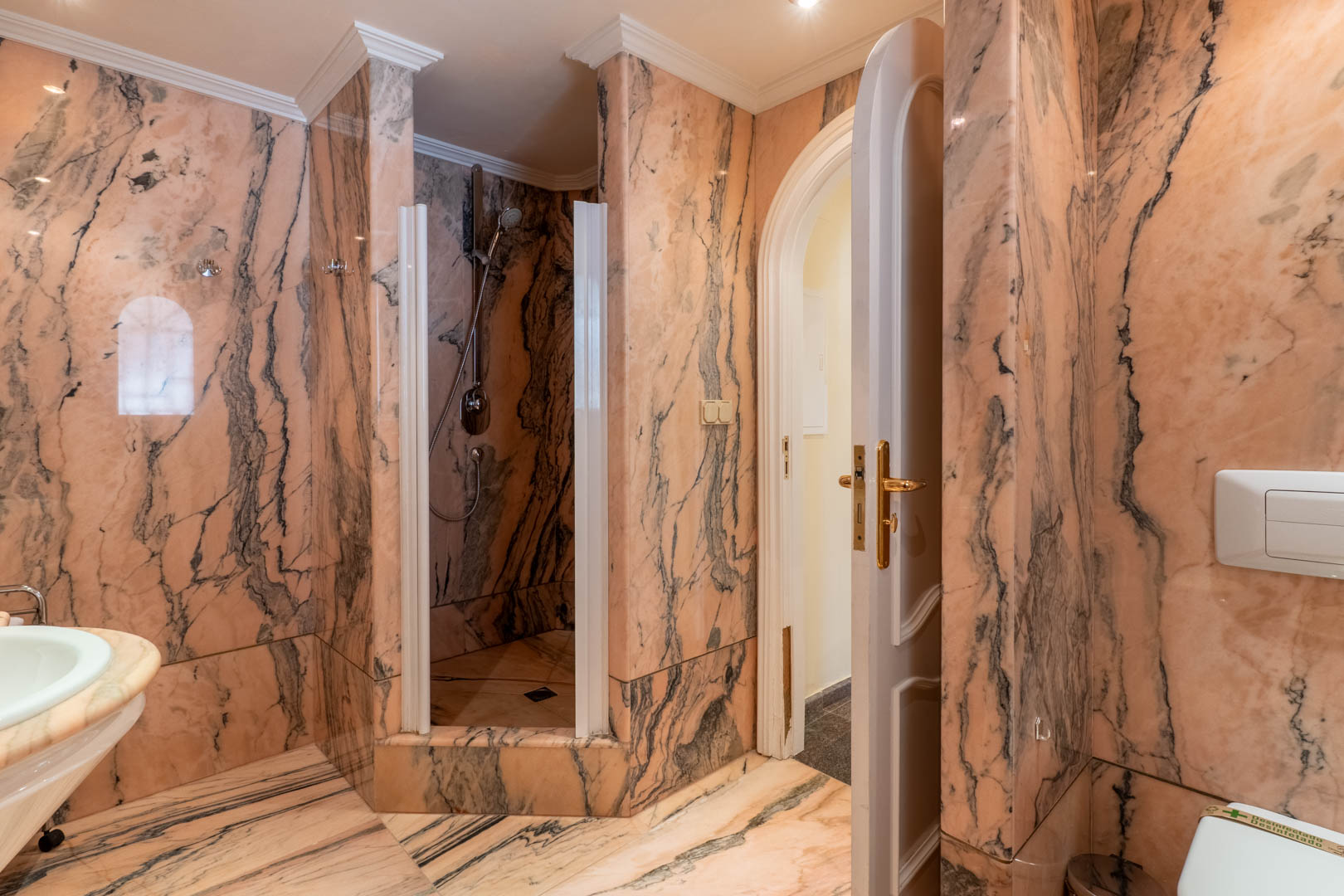 Badezimmer mit vollständiger rosa Marmorverkleidung, einer begehbaren Dusche, klassischen Deckenleisten und einer Rundbogentür mit goldenen Beschlägen.
